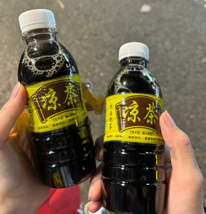Two plastic bottles filled with dark-colored traditional Chinese herbal tea, featuring yellow labels with the Chinese characters for "Liangcha," held by a person against a blurry outdoor background.