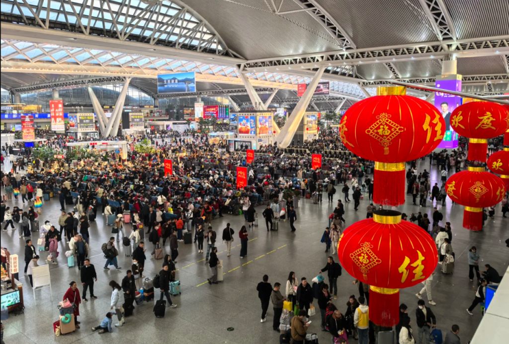 A vast, crowded departure hall of a modern Chinese railway station during the Spring Festival travel rush, with large decorative red lanterns in the foreground and thousands of travelers with luggage.