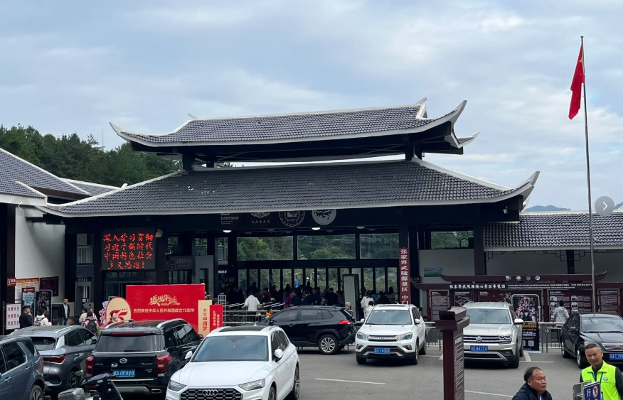 The main entrance gate to the Zhangjiajie Wulingyuan Scenic Area, featuring traditional Chinese architecture with a double-tiered grey tiled roof. The facade displays UNESCO and 5A tourist attraction logos. Tourists are queuing at the turnstiles, and the foreground is filled with parked cars, with a Chinese flag flying on the right.